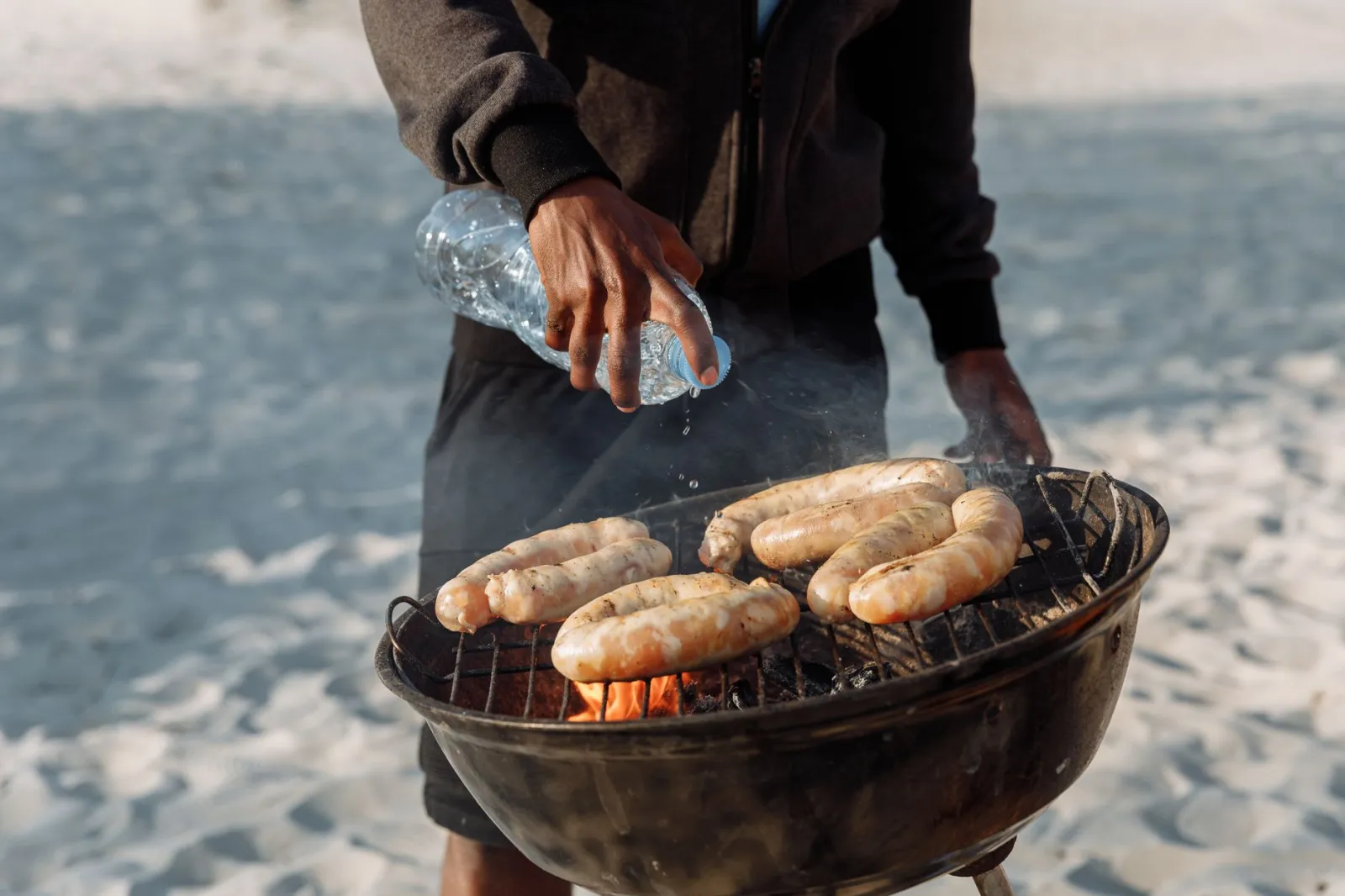 Person grilling sausages on a beach with a portable barbecue and bottled water.