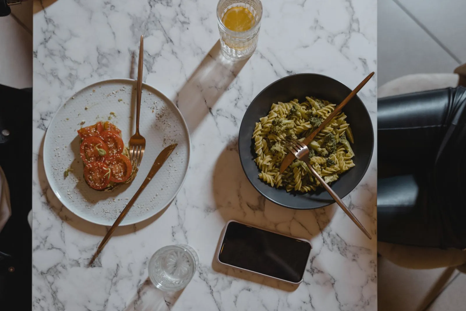 Overhead view of a vegan pasta and tomato salad meal on a marble table with a phone.